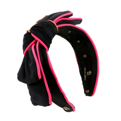 A black neoprene wide headband with a knotted bow on the front, trimmed with bright pink piping and small gold decorative details, shown on a plain white background.